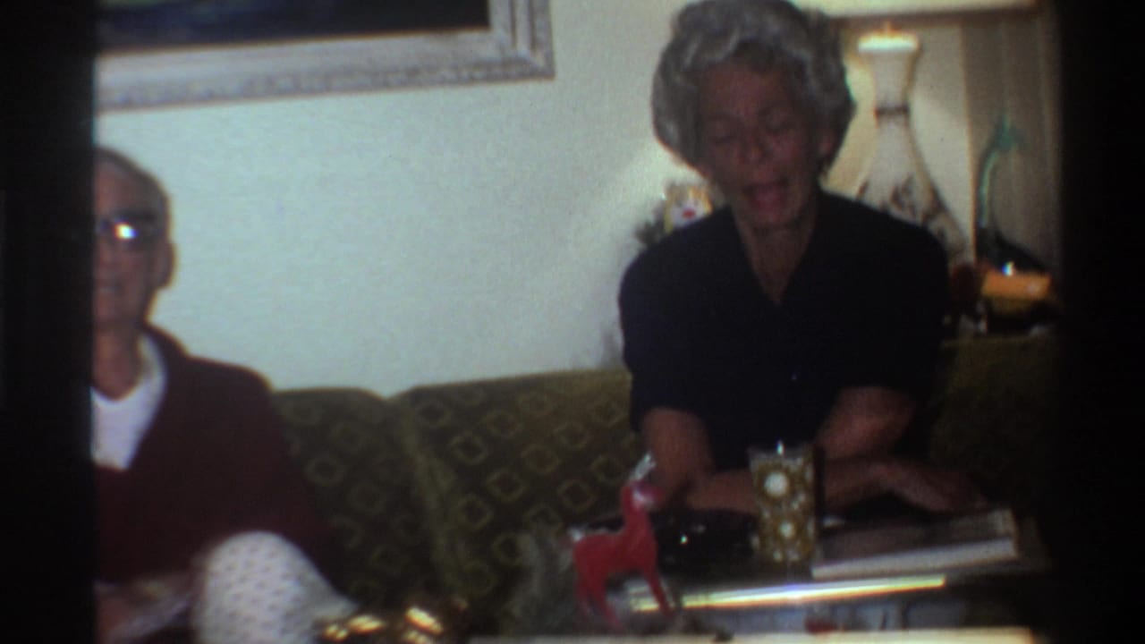 Elderly man in dark red sweater and older woman with gray hair seated on patterned couch mid-conversation, Christmas gifts with gold bow visible in foreground.