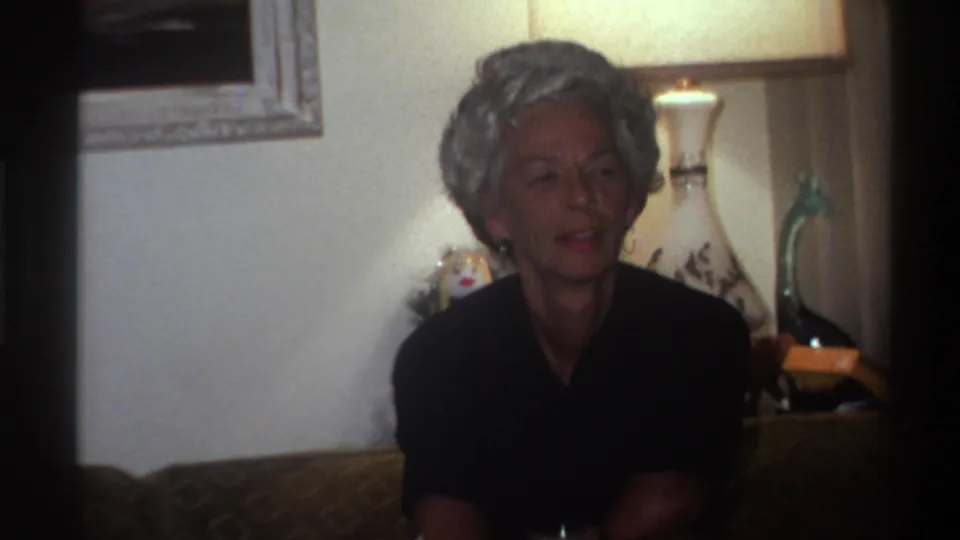 1975 Gainesville, Florida — close view of older woman with styled gray hair in dark top appearing to speak, white decorative lamp and framed landscape painting behind her, 8mm home movie.
