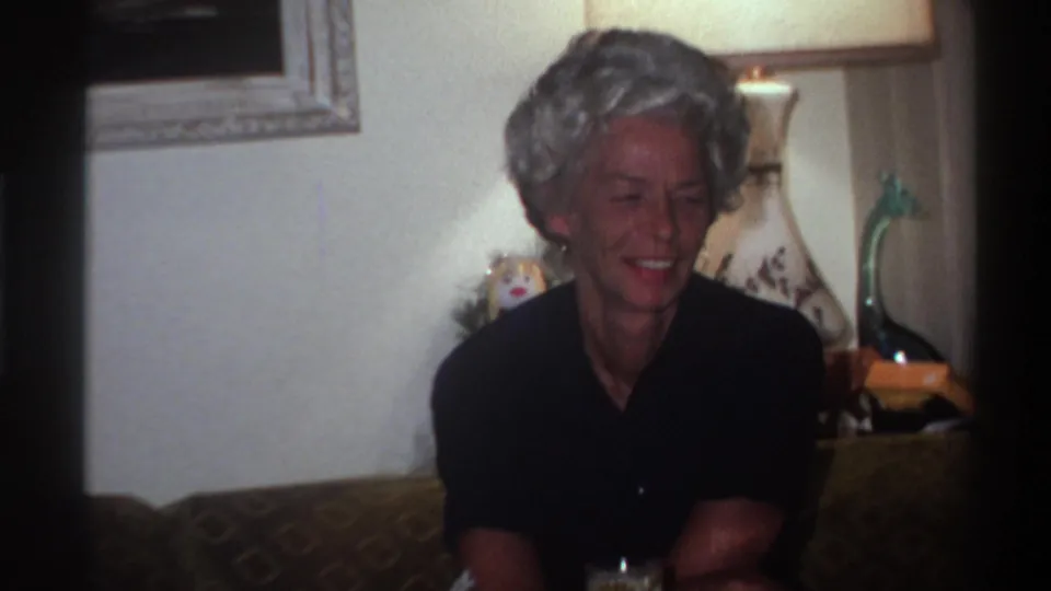 1975 Gainesville, Florida — close view of older woman with styled gray hair smiling broadly during Christmas family gathering, white decorative lamp and painting behind her, 8mm home movie.