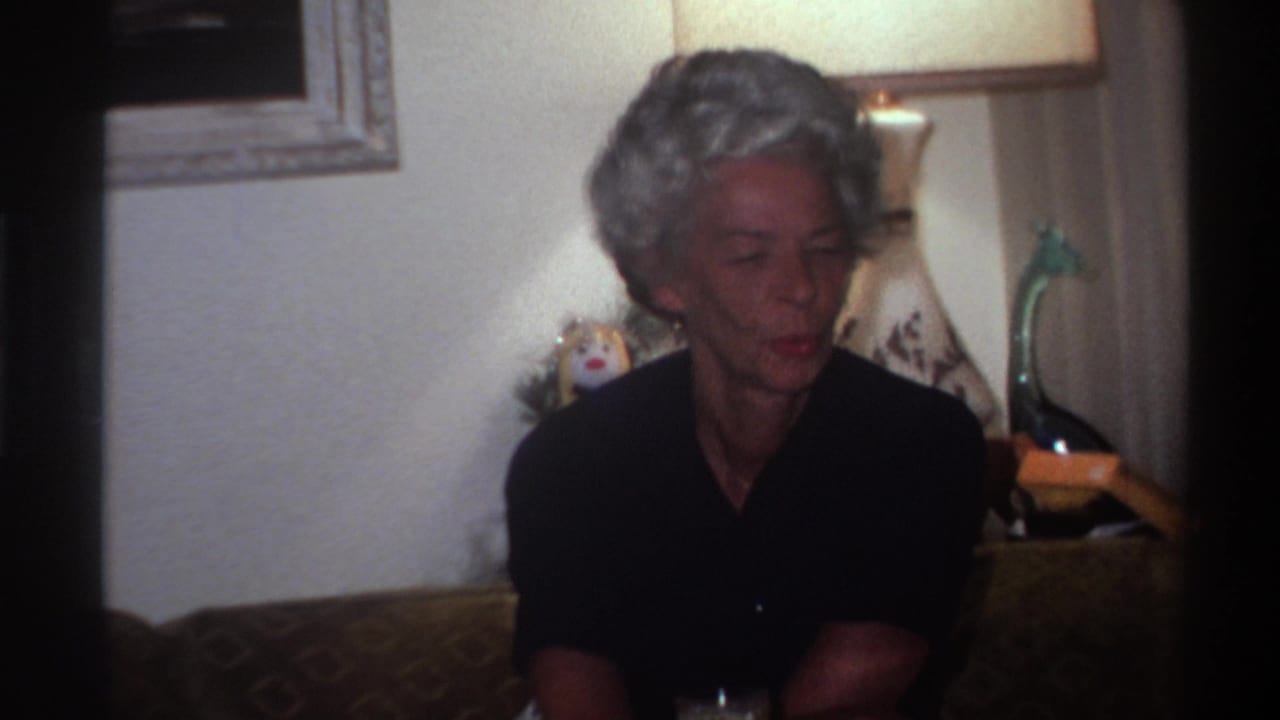 Elderly woman with short gray hair in dark top, seated on patterned upholstered sofa in a warmly lit living room interior, circa 1975 Florida.