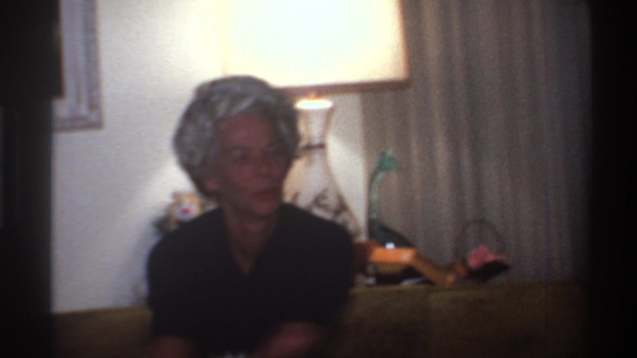 Elderly woman with short gray hair in dark top, seated in a 1970s warmly lit interior with table lamp and decorative items visible behind her.