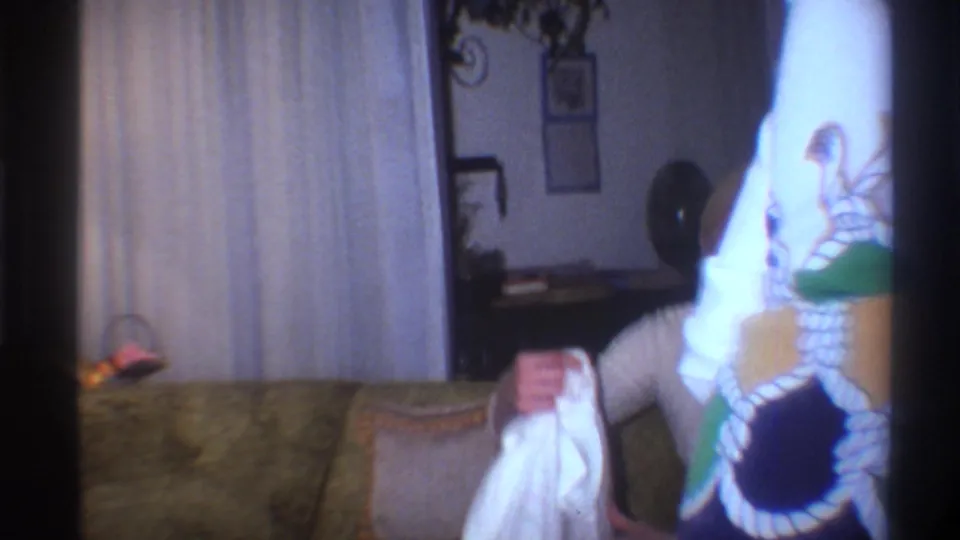 Close view of hands folding white laundry in a 1970s interior; large floral-print fabric, sheer curtains, and a small wall ornament visible in background.