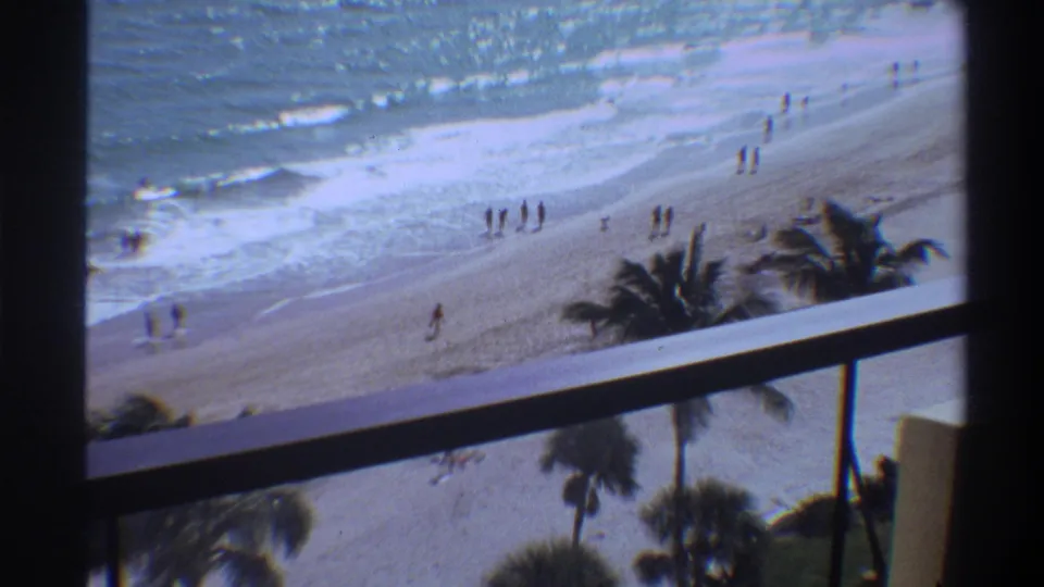 Elevated balcony view of a Florida beach circa 1975, with palm trees in foreground, rolling ocean waves, and scattered beachgoers walking along the sand.
