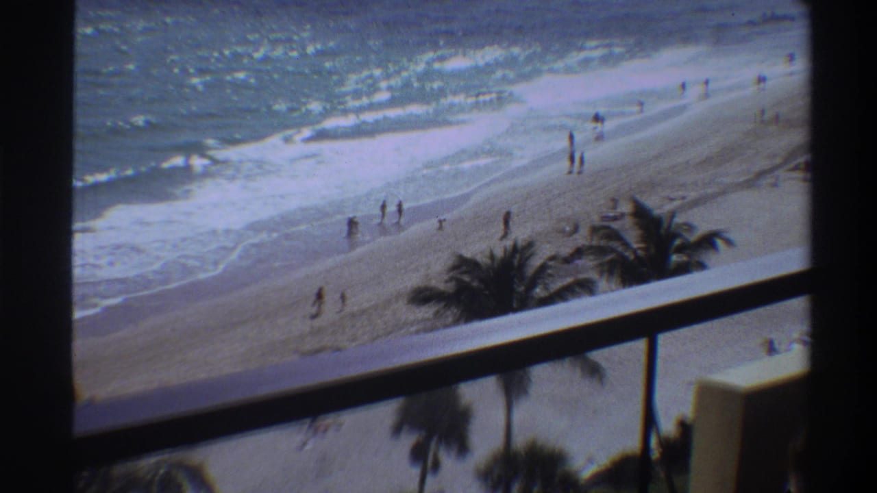 8mm frame circa 1975 showing a Florida beach from an elevated balcony; scattered beachgoers near breaking waves under palm trees.