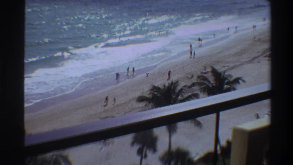8mm frame circa 1975 showing a Florida beach from an elevated balcony; scattered beachgoers near breaking waves under palm trees.