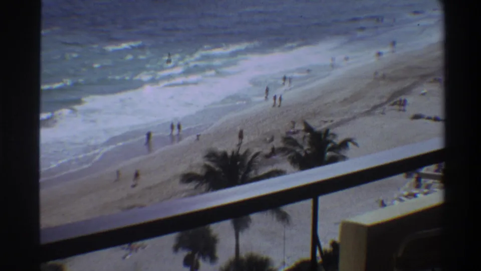 Mid-1970s 8mm home movie frame of a Florida beach from an elevated balcony; palm trees, rolling surf, and small distant figures on a wide sandy shore.