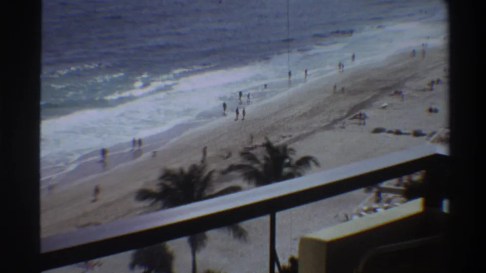 Final frame of a 1975 Florida beach home movie; balcony perspective shows palm trees, surf, and small distant figures on a wide sandy shore.