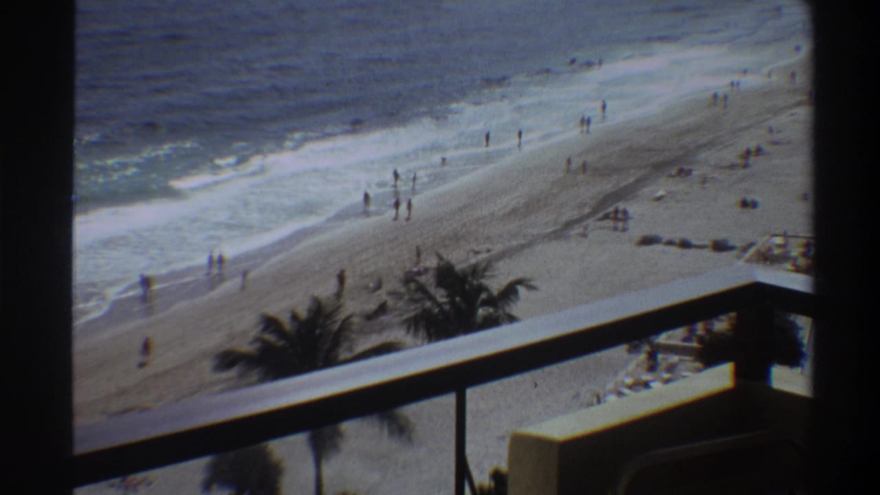8mm home movie shot from a hotel balcony overlooking a Florida beach, circa 1975, with palm trees in foreground and distant beachgoers.