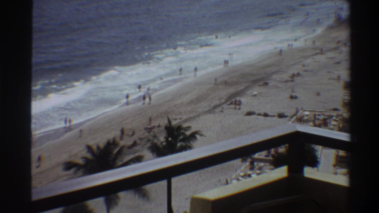 Balcony-height view of a Florida beachfront, circa 1975, showing beachgoers on sand and at the water's edge with rolling waves and palm trees.