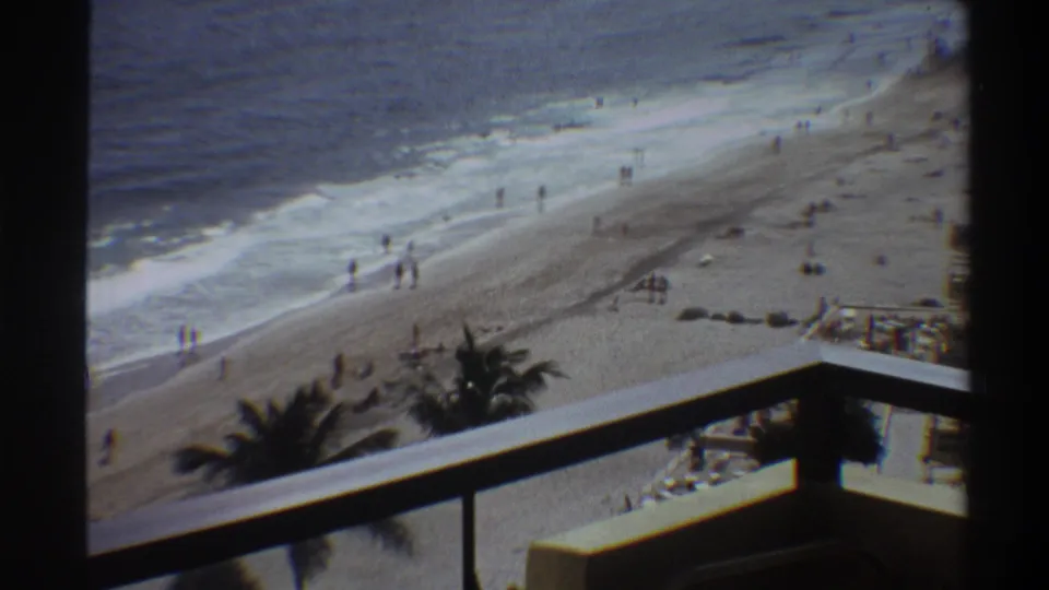 Balcony-height view of a Florida beachfront, circa 1975, showing beachgoers on sand and at the water's edge with rolling waves and palm trees.
