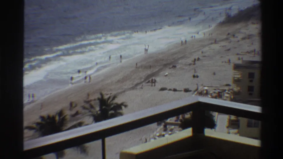 Hotel or resort balcony vantage point over a Florida beach, circa 1975; building roofline visible below, beachgoers on sand and at the wave line.