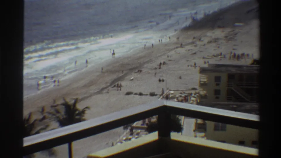 Final frame of a 1975 Florida beach home movie: balcony railing foreground, hotel building below, beach items and figures on the sand near calm surf.