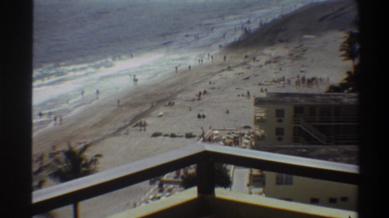 High balcony view looking down at a wide sandy beach, with a multi-story beachfront building below and breaking waves along the shoreline.