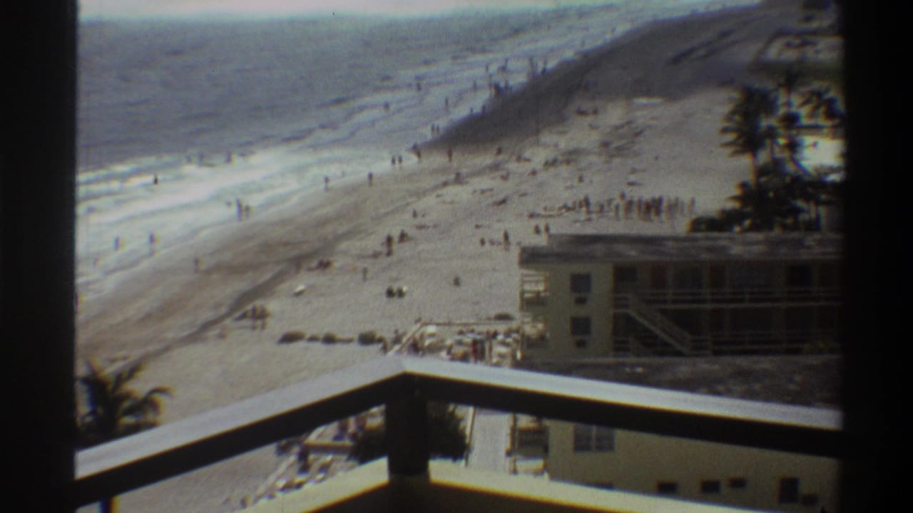8mm home movie circa 1975, Florida coast: veranda POV showing hotel facade, wide sandy beach with scattered bathers, palm trees, and ocean waves.