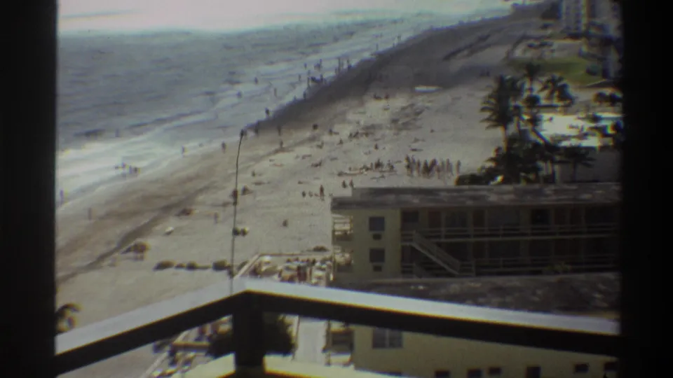 8mm home movie circa 1975, Florida coast: elevated balcony view with palm trees prominent in the mid-ground, hotel with external staircase below, and crowds on the beach.