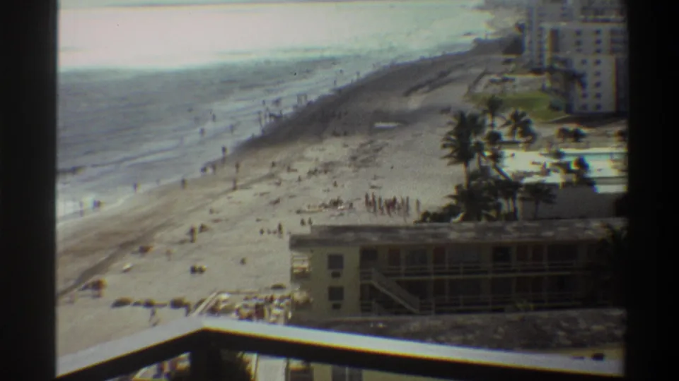 8mm home movie circa 1975, Florida coast: wide beach overview from high balcony, beachfront building below, a second taller structure visible in the distance, and scattered beachgoers.