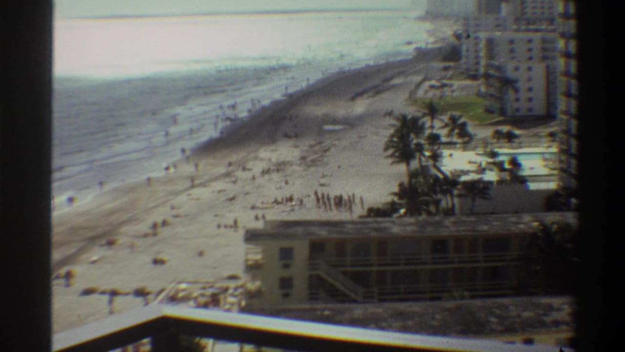 High-angle 8mm view of a Florida beachfront resort strip, circa 1975, with wide sandy shore, scattered beachgoers, palm trees, and a multi-story hotel skyline.