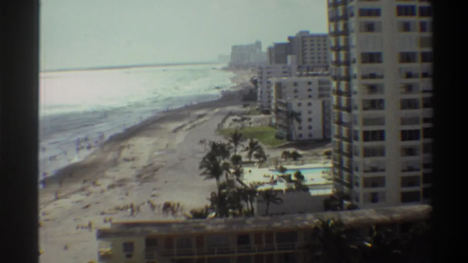 Continuation of 8mm pan across a Florida beachfront resort, circa 1975, with high-rise hotels, palm trees, and a pool area in the foreground.