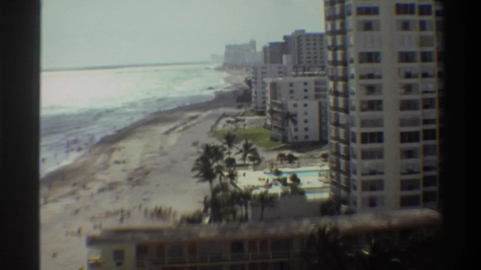 High vantage-point 8mm home movie circa 1975 of a Florida coastal resort, showing high-rise apartment towers, palm trees, a pool, and beach.