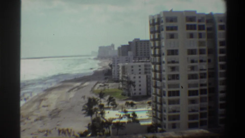Closer 8mm frame circa 1975 of a multi-storey beachfront apartment tower with a palm-lined pool area and surf along a Florida coast.
