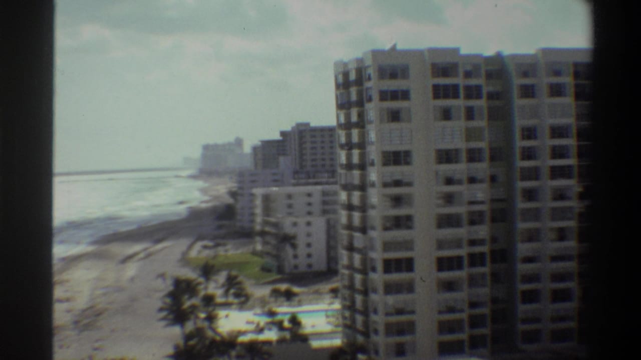 8mm home movie ca. 1975: elevated view of Florida beachfront high-rise towers, sandy shore, palm trees, swimming pool, and ocean horizon under overcast sky.