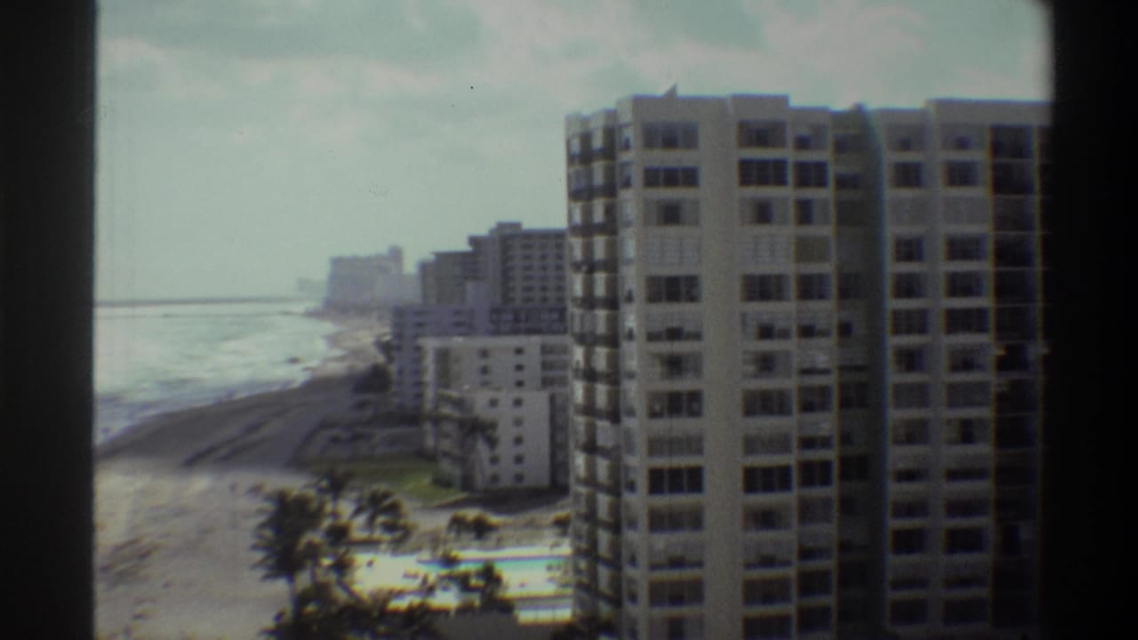 Elevated 8mm view of beachfront high-rise towers with ocean behind, palm trees lower-left, and a pool visible under an overcast sky.