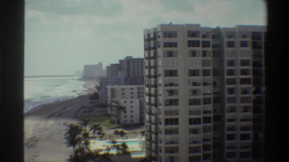 8mm frame ca. 1975: Florida coastal high-rise complex viewed from elevation, with ocean surf, palm trees, and pool visible in the lower foreground.