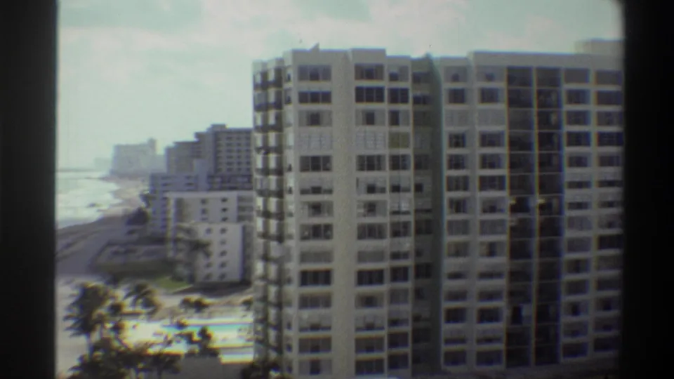 8mm home movie ca. 1975: a Florida beachfront high-rise dominates the foreground as ocean surf, sandy beach, and palm trees appear to the left.