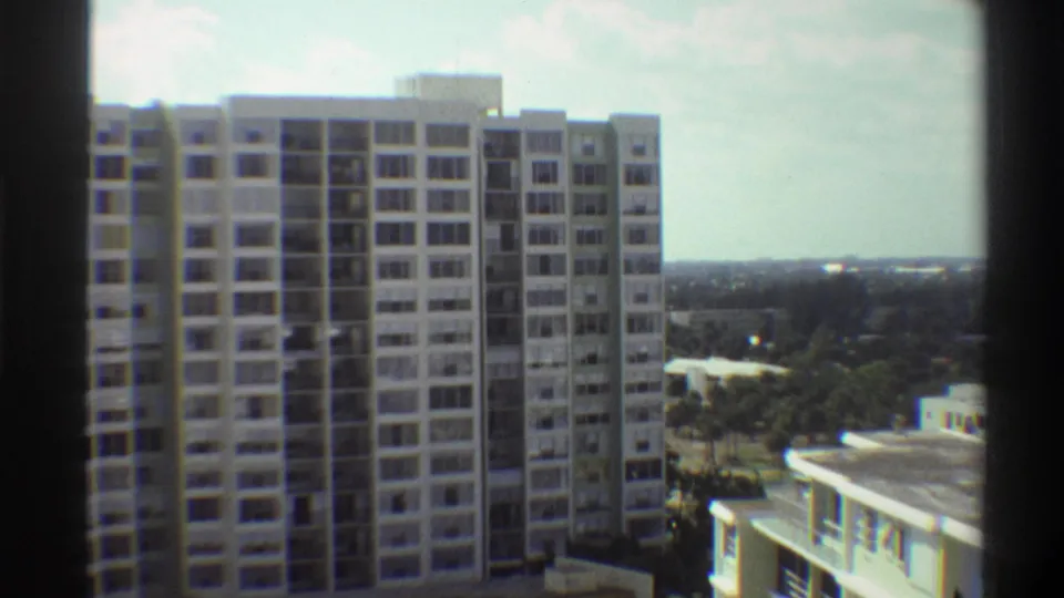 8mm home movie circa 1975, elevated view of a tall Gainesville, Florida apartment block with a flat-roofed lower building in the foreground and a distant treeline visible.