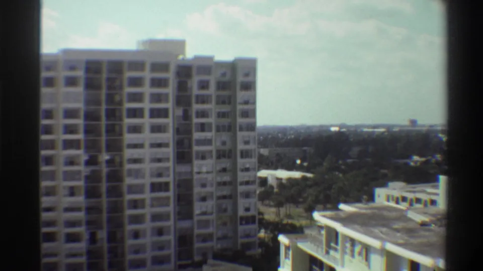 Final 8mm frame circa 1975 showing the full apartment tower centred against a broad Florida treeline and pale sky, captured from a high residential vantage point.