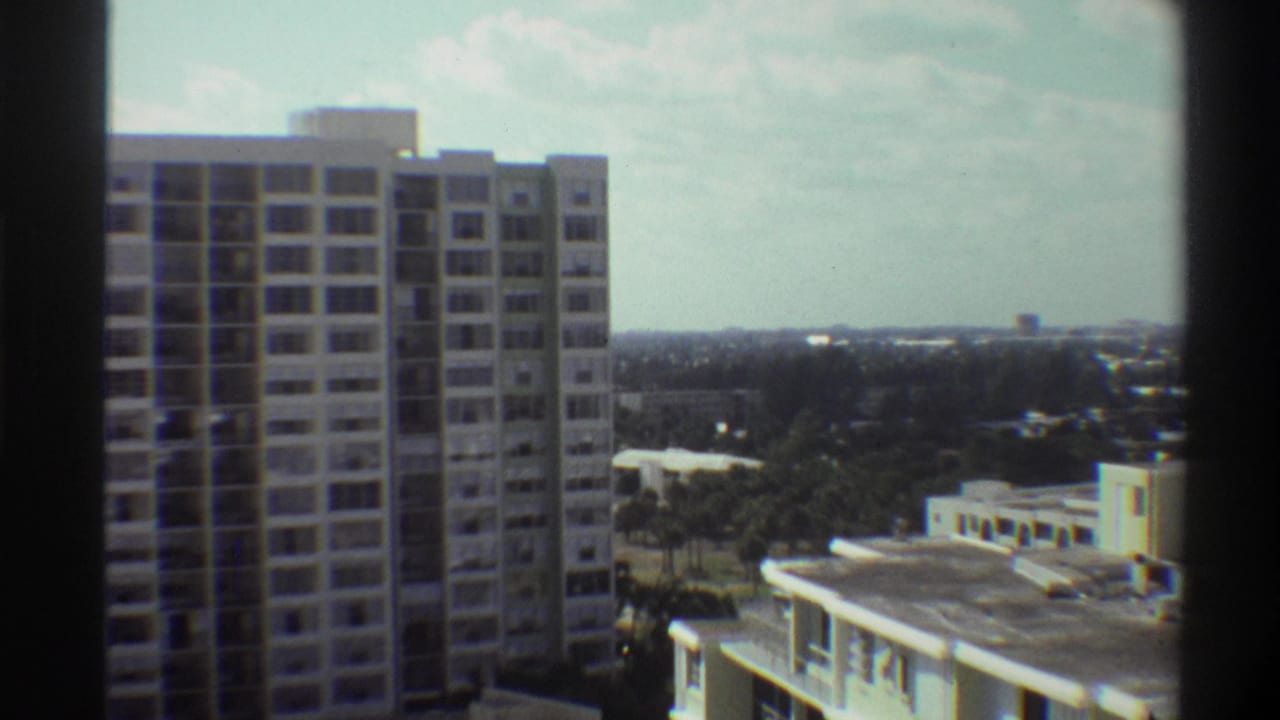 Super 8 frame from 1975 showing a high-rise residential tower and flat-roofed mid-rise in Gainesville, Florida, with subtropical tree canopy and overcast sky.