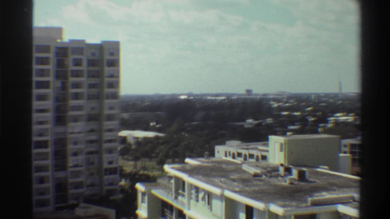 Mid-pan Super 8 frame from 1975 Gainesville, Florida: concrete tower at left, flat-roofed buildings, and a wide subtropical landscape under overcast sky.
