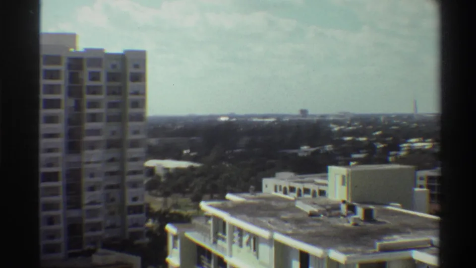 Mid-pan Super 8 frame from 1975 Gainesville, Florida: concrete tower at left, flat-roofed buildings, and a wide subtropical landscape under overcast sky.