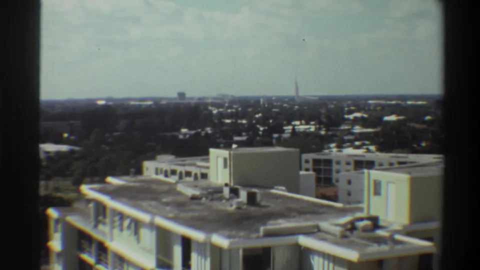 Final pan frame in Super 8 from 1975 Gainesville, Florida: flat rooftops, subtropical tree cover, and wide overcast horizon; tall tower now out of frame.