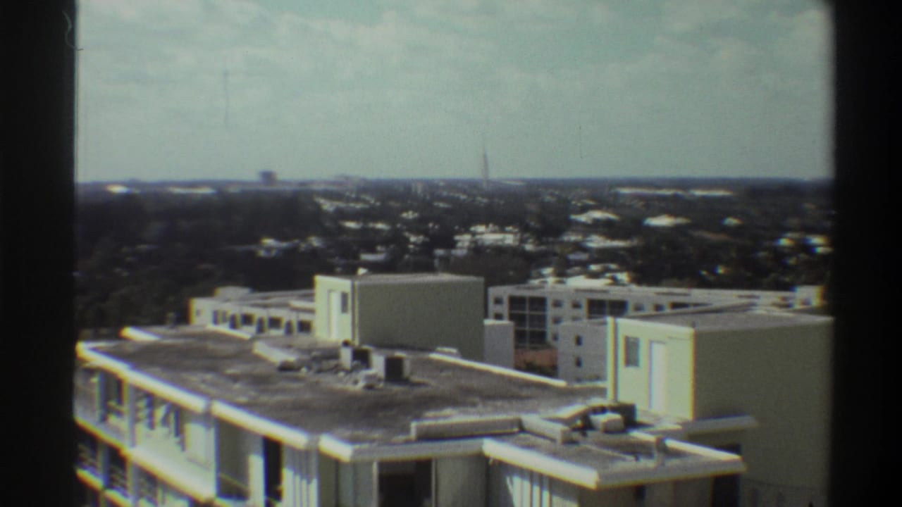 High vantage point over mid-century modernist buildings in what appears to be Gainesville, Florida, circa 1975, with suburban sprawl and a distant spire.