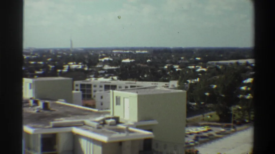 Wider pan reveals tree-covered suburban sprawl in what appears to be Gainesville, Florida, circa 1975, with an institutional rooftop and parking lot in the foreground.