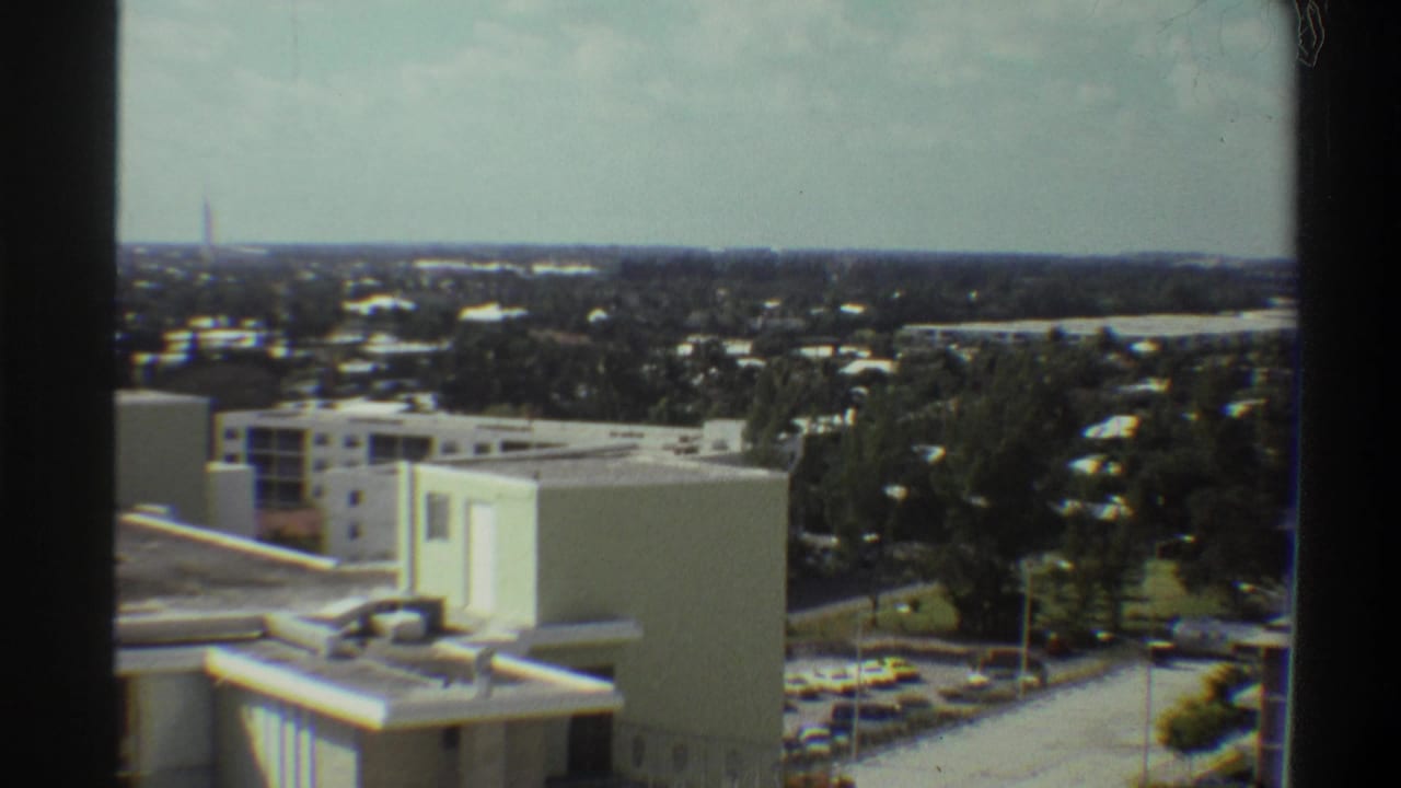 8mm footage circa 1975 showing an elevated window view over a flat-roofed building, surface parking, and subtropical tree canopy in a Florida suburb.