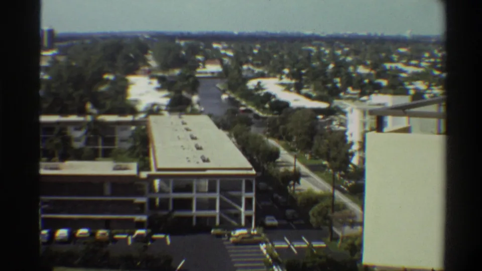 8mm footage circa 1975 showing a multi-level open structure, wide road, yellow vehicles, and low-rise Florida suburban development spreading to a flat horizon.