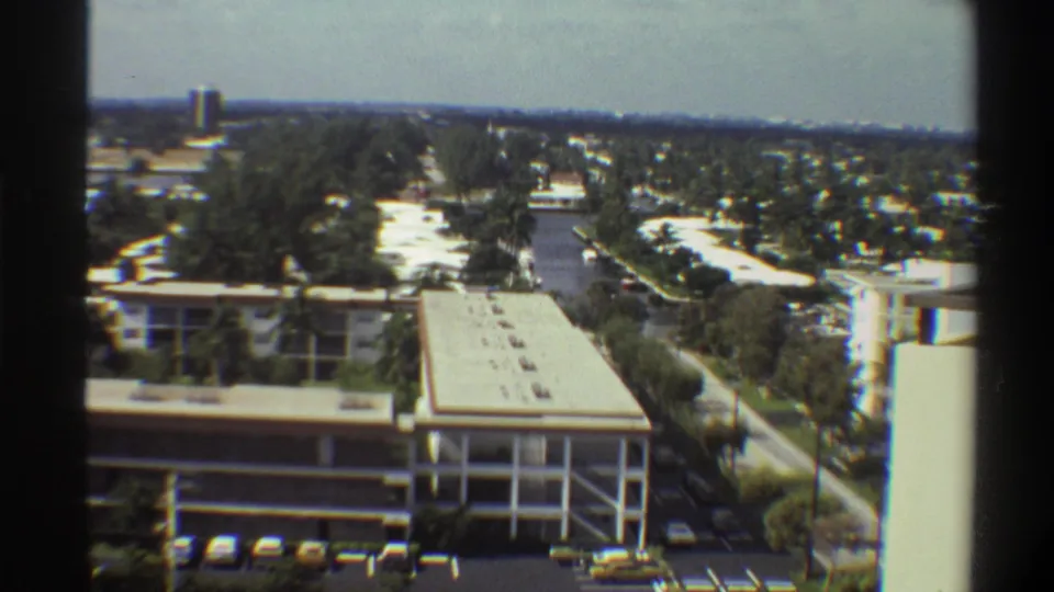 8mm footage circa 1975 showing an open-bay multi-story structure, tree-lined street, yellow vehicles, and low-rise buildings across a flat Florida suburban landscape.