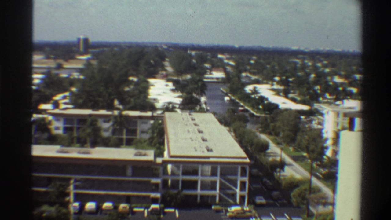 Elevated 8mm film view of a 1970s flat-roofed institutional building in Gainesville, Florida, with tree canopy and distant horizon.