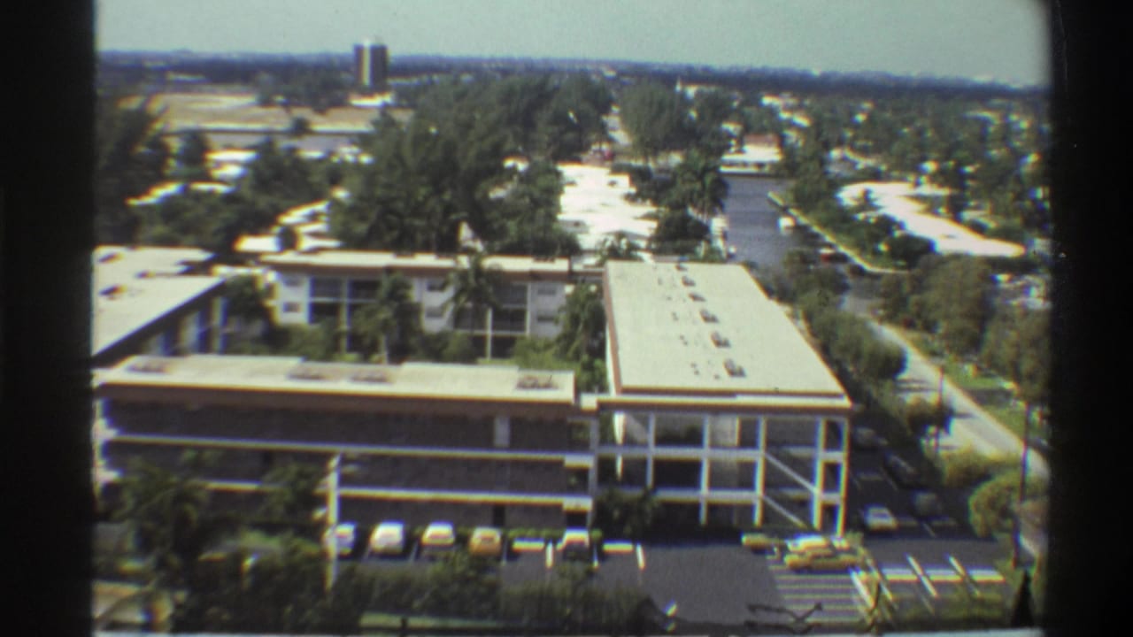Elevated pan view with flat-roofed building reframed right, parking area visible, dense tree canopy filling midground.