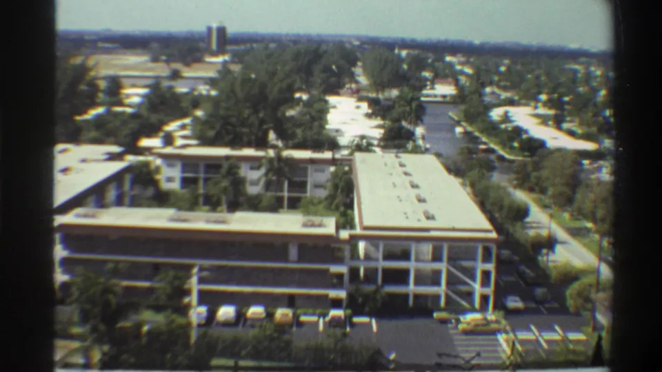 Mid-pan 8mm frame of a 1970s institutional complex in Gainesville, Florida, with parking lot foreground and distant suburban horizon.