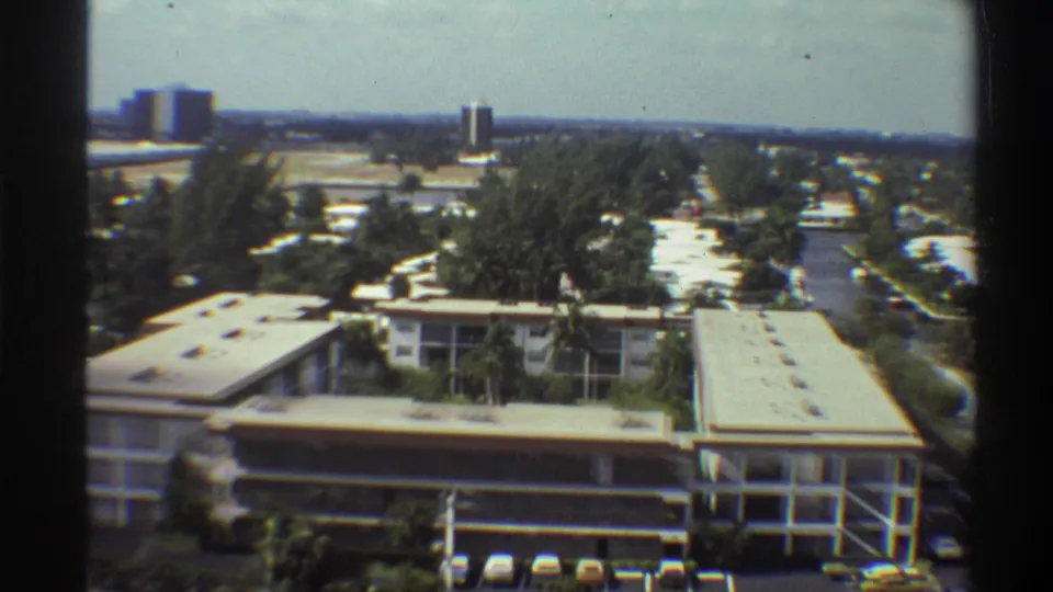 Elevated 8mm pan view of 1970s Gainesville, Florida, showing institutional building, tree canopy, and distant high-rise towers.