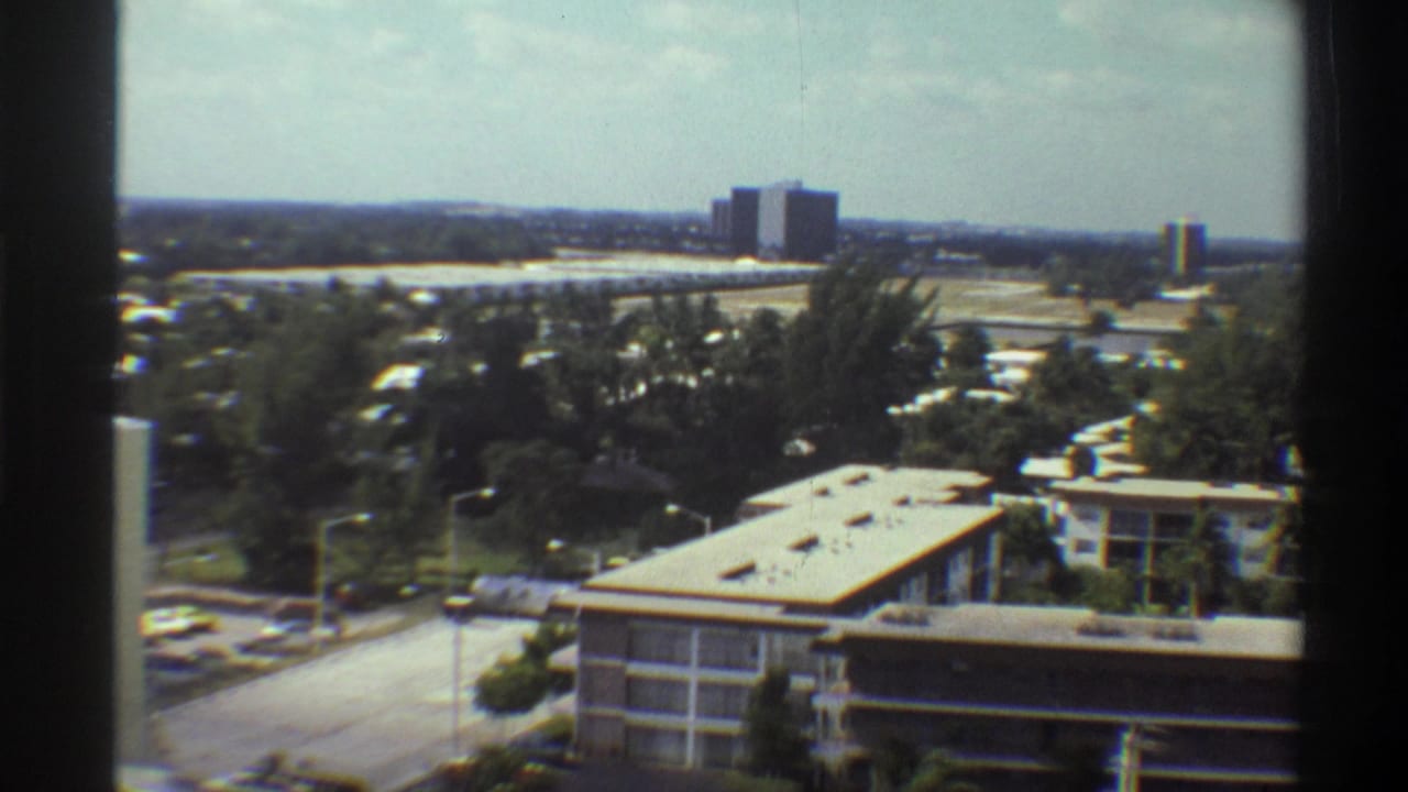 Elevated 8mm view over a Florida suburban neighborhood, circa 1975, showing a flat-roofed low-rise building, parking area, dense trees, and a distant skyline consistent with Gainesville.