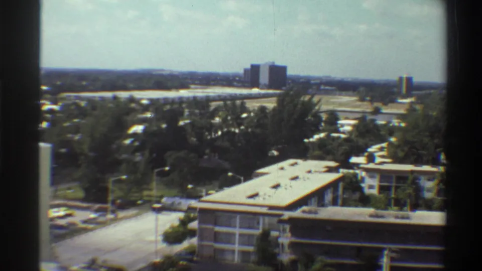 Elevated 8mm view over a Florida suburban neighborhood, circa 1975, showing a flat-roofed low-rise building, parking area, dense trees, and a distant skyline consistent with Gainesville.