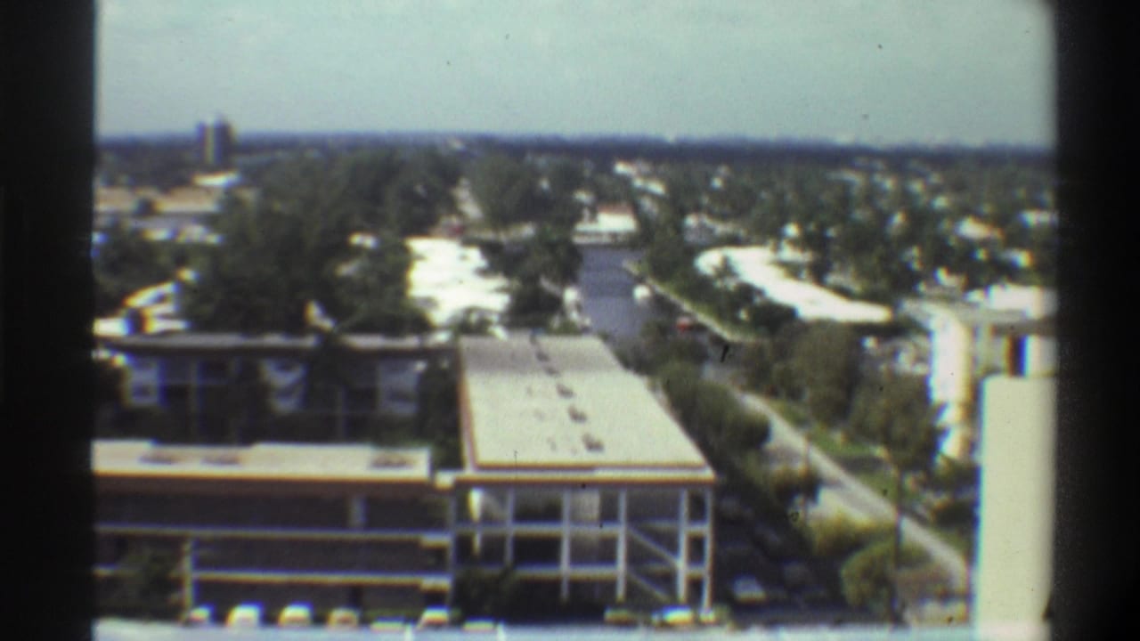 Mid-pan 8mm frame over what appears to be Gainesville, Florida, 1975, showing low-rise architecture, a roadway running alongside the building, and dense Florida vegetation beyond.