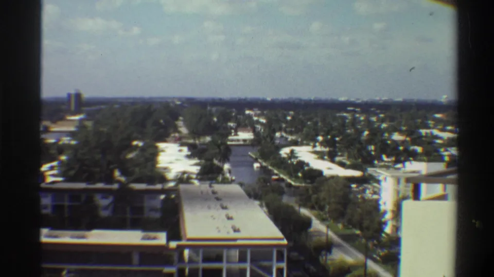Wide 8mm panorama over Gainesville, Florida suburbs circa 1975, showing flat terrain, palm trees, low-rise rooftops, and a possible canal or waterway cutting through the middle ground.
