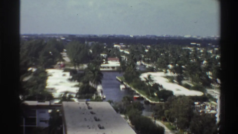 Final pan frame over Gainesville, Florida, 1975, showing a water channel or canal among palm trees and low-rise suburban buildings under an overcast sky.