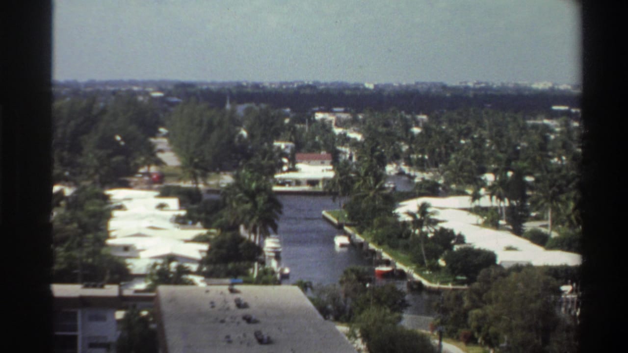 High-angle 8mm shot of a Florida canal residential community, circa 1975 — palm trees, low-rise flat-roofed homes, and boats along the waterway under a hazy sky.