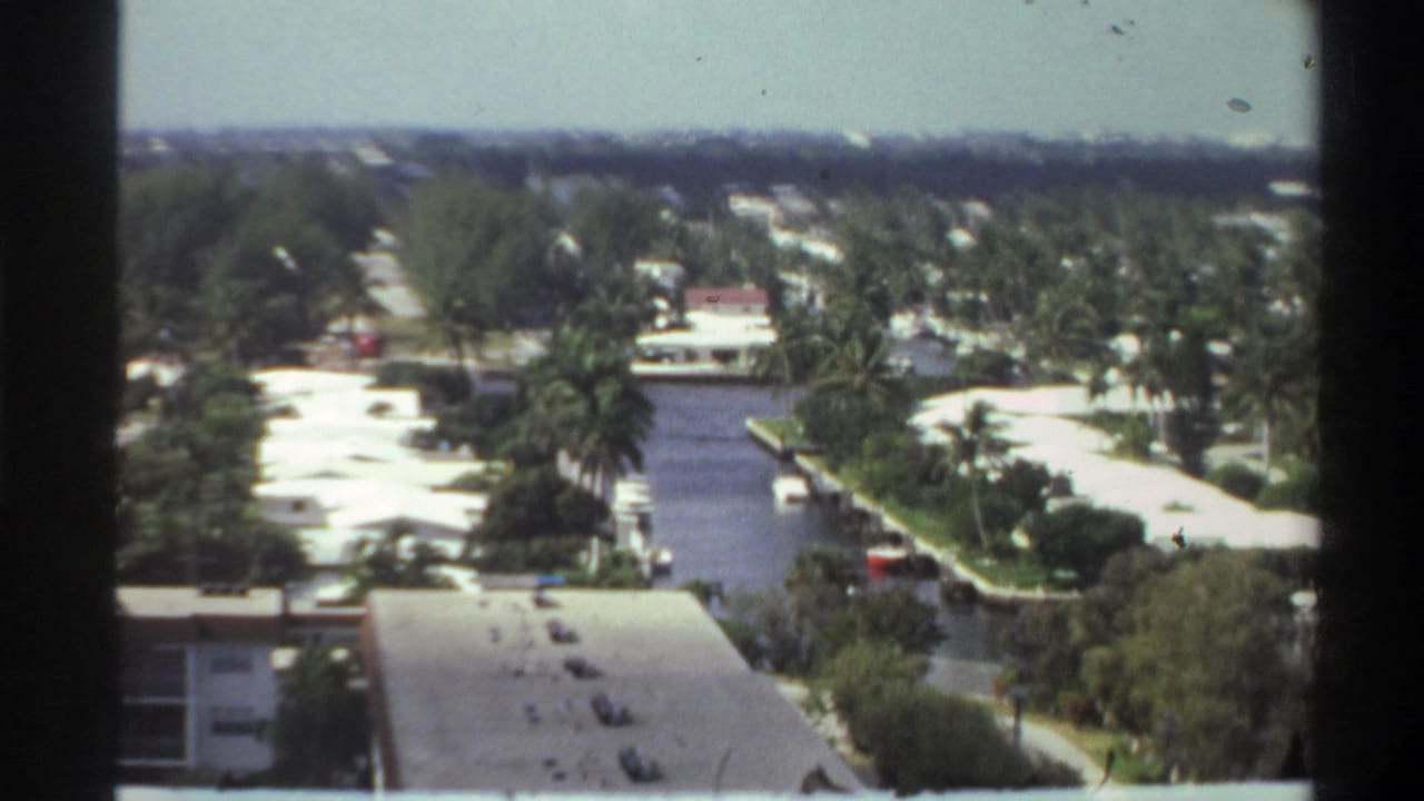 Continuation of high-angle 8mm shot over a Florida canal community, circa 1975 — waterways, palm canopy, low-rise residential rooftops, and a boat visible along the canal.